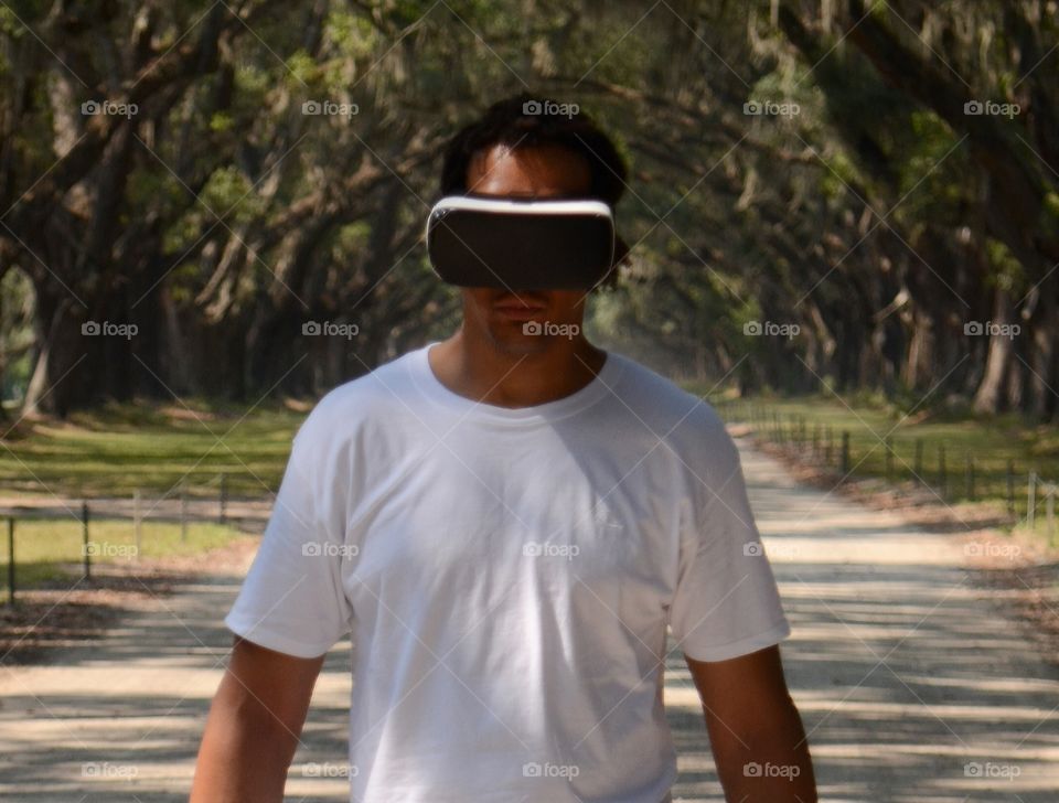 A young man wearing a virtual reality mask is transported to the picturesque road lined with more than four hundred live oak trees that over hang Oak avenue which lead to the heart of Wormsloe State Historic Site and plantation in Savannah, Georgia.