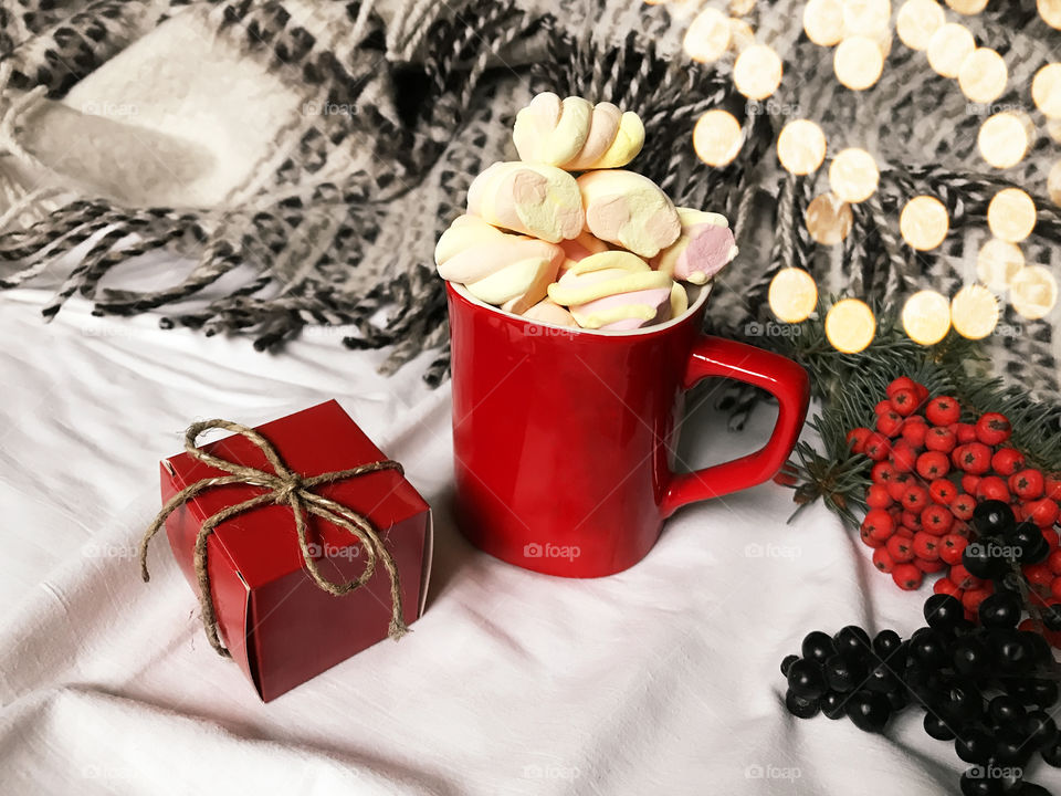 Cup of hot drink with marshmallows , gift box and branch of Christmas tree with lights in cozy bed with blanket 