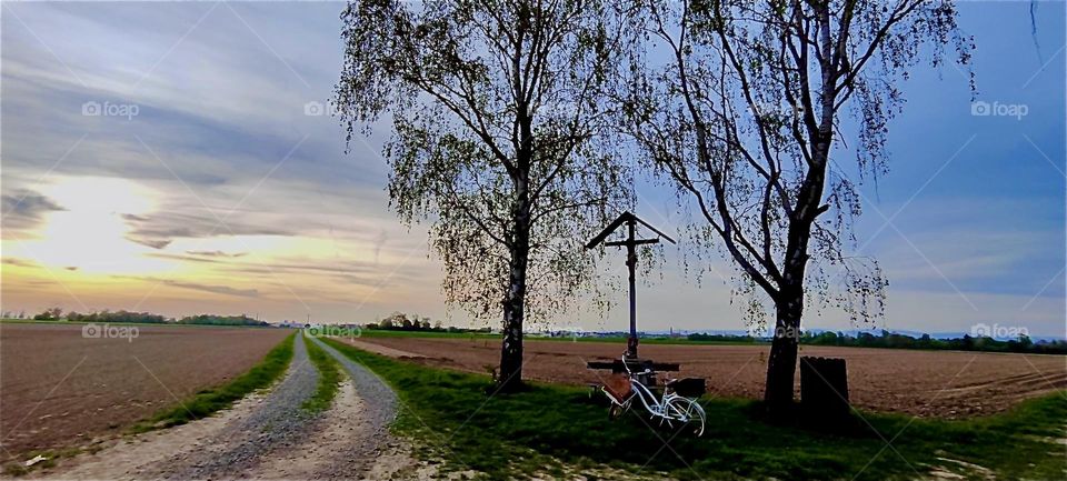 The country road or trail for bicyclists and pedestrians stretches endlessly into the distance in rural “Bavaria”, Germany. There is a rest stop with a bench and a crucifix underneath a symbolic triangle roof. 2024. Hypnotic Productions
