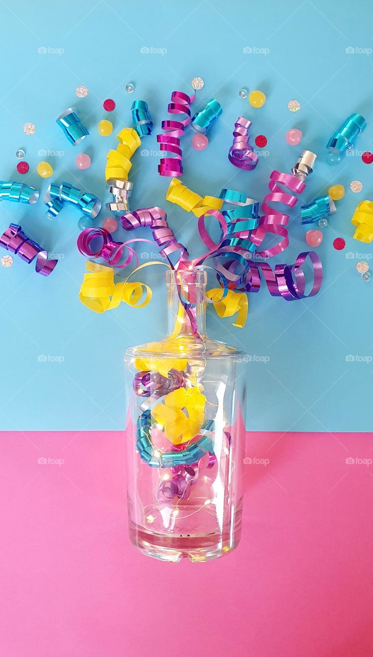 Colour explosion flatlay on blue and pink background composed of pink, blue, yellow, purple and silver gift package ribbons placed as if exploding out of a clear bottle