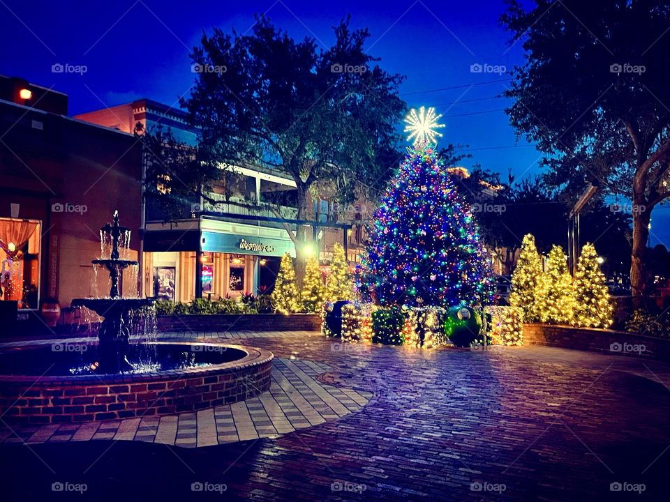 Holiday lighting in a quaint town square at night with a fountain and shops in the background 