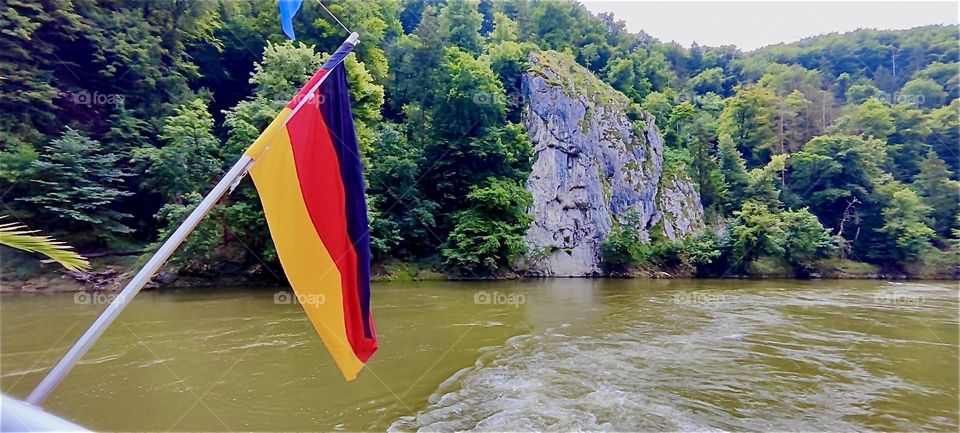 The black red & gold German flag is hoisted at the bow of a “Zille”, a large fishing row boat making its way along the “Danube” between “Kelheim” and the “Weltenburg Abbey” at the “Donaudurchbruch” or “Danube Gorge”. 2024. Hypnotic Productions