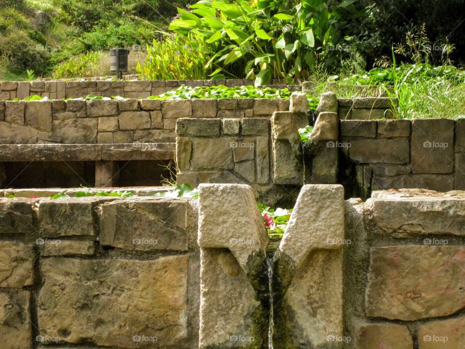 Artificial stream with stone terraces, green plants and part of a pink flower