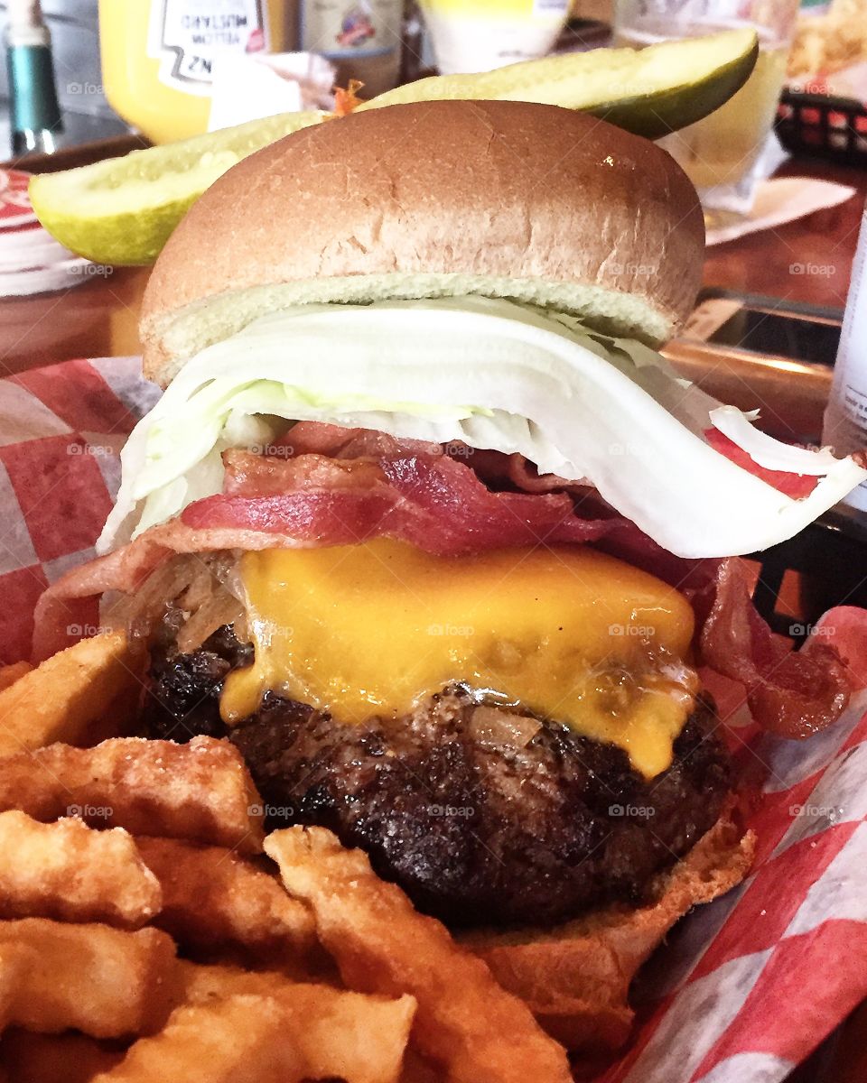 Big, juicy bacon cheeseburger in a basket with crinkle cut fries and a dill pickle slice on top.