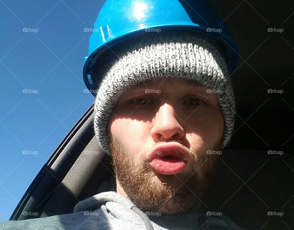 Work face, man in a construction hat with a beanie making a funny face. He has charming blue eyes.