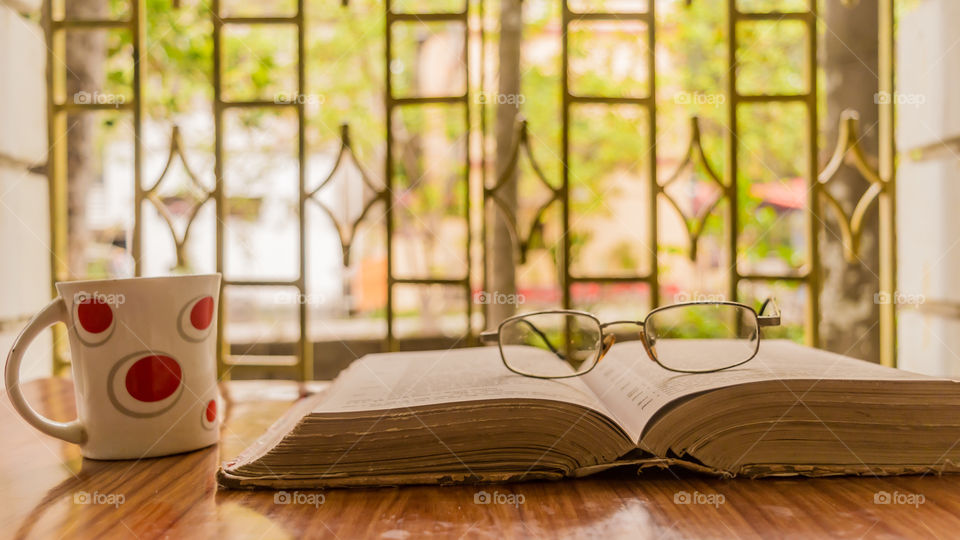 An open book a coffee cup and a specs or eyeglass on wooden table, warm sunny morning, side view close up. Education or Holiday Concept. Isolated background in selective focus, shallow depth of field