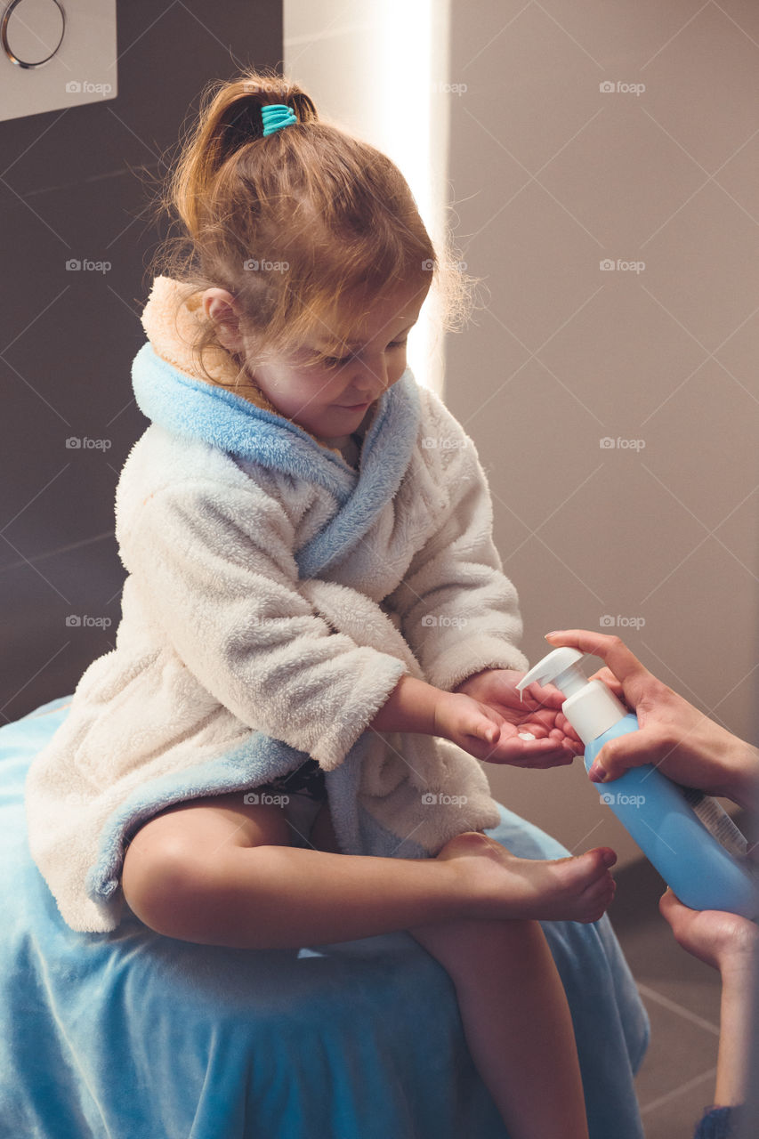 Mother applying moisturizing cream on her daughter's legs after bath. Mom caring about her child. Girl sitting in bathroom, wearing bathrobe. Real people, authentic situations