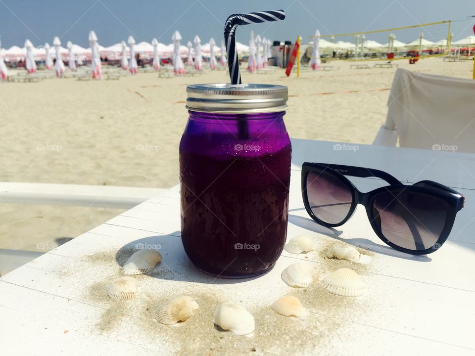 Jar of blueberries lemonade on white wooden table surrounded by seashells and pair of black sunglasses