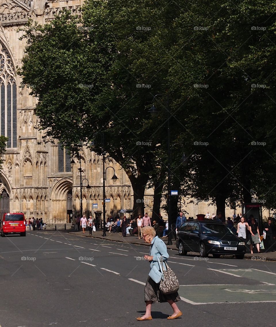 A lady hurriedly crosses a street with a cathedral in the background on a sunny summer day in England. 