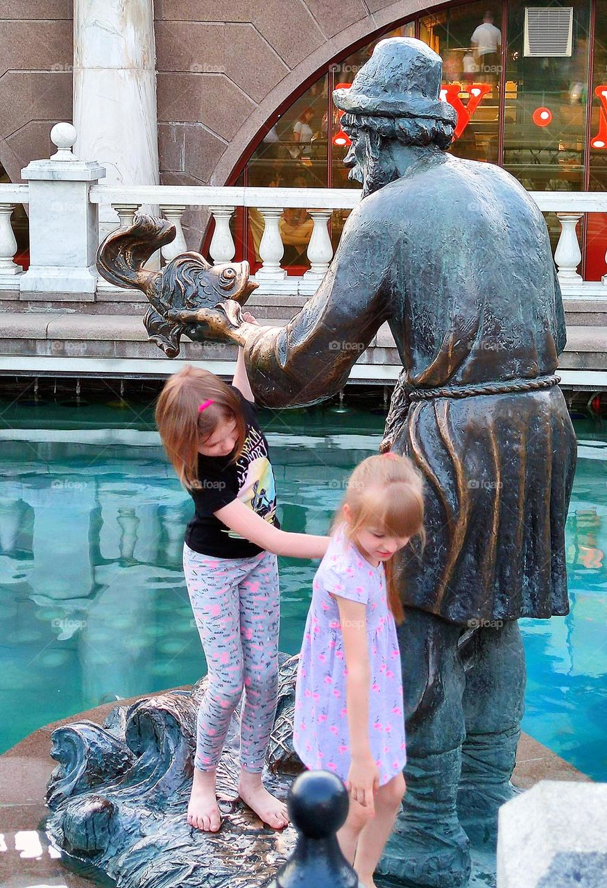 Summer in the city. Two little girls descend from the sculpture of the fountain: "The old man and the goldfish"