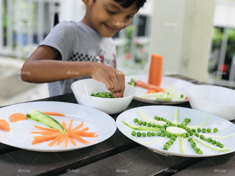 Kids having fun by doing creative activities with vegetables in summer time.A 6 year old boy making sun , tree with green peas and carrots cucumber in summertime as a activity.