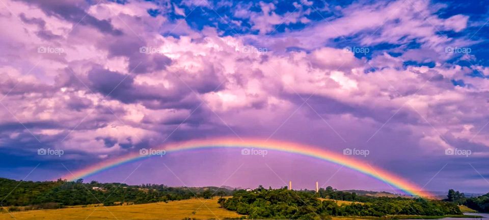 Landscape with beautiful Rainbow and clouds with pink and purple tones, contrasting with the blue sky.
Paisagem com lindo Arco-íris e nuvens com tonalidade rosa e roxo, contrastando com o céu azul.