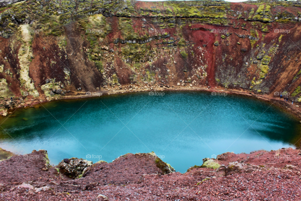Edge of the overlook of Kerid crater in Iceland. The water is naturally an electric blue color with bright red soil surrounding the crater