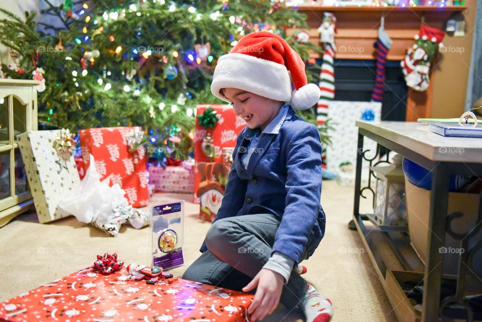 Smiling young boy wearing a Santa hat and opening a Christmas present in a decorated home