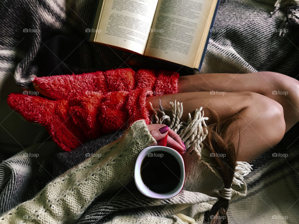 Young woman in pink home boots reading a book and drinking coffee in cozy bed 