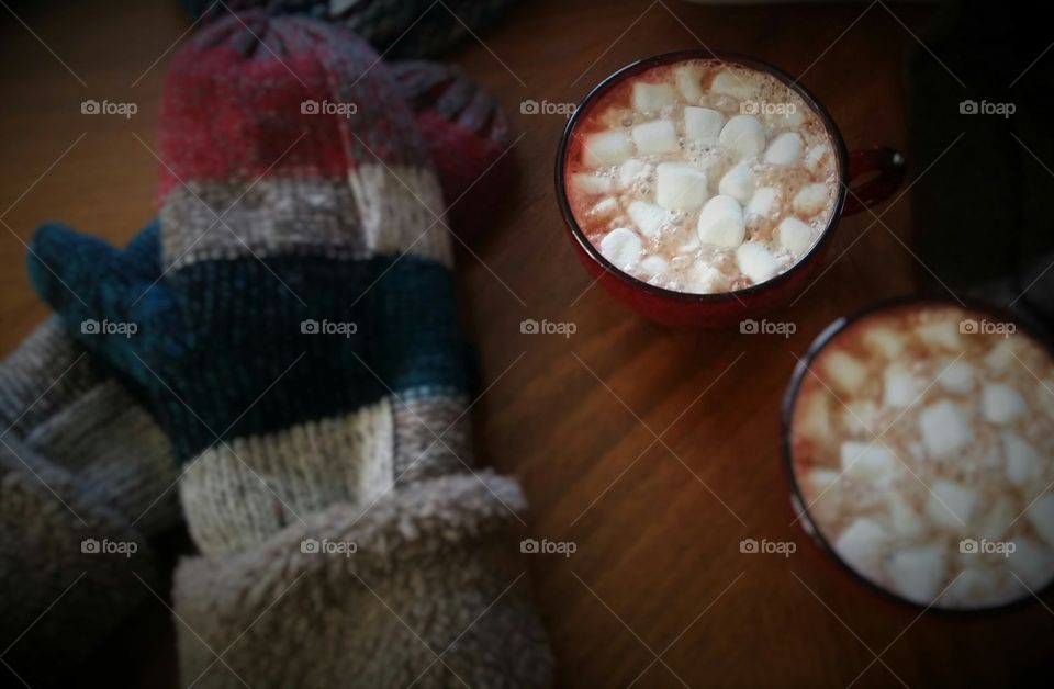 A pair of mittens sitting with two cups of hot cocoa with melting marshmallows on a wooden table