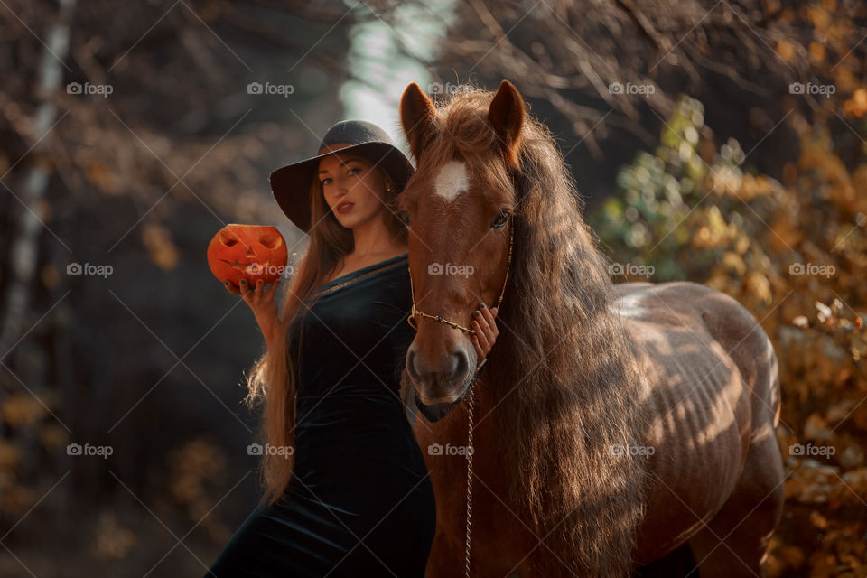 Woman in witch wear with horse in autumn park 