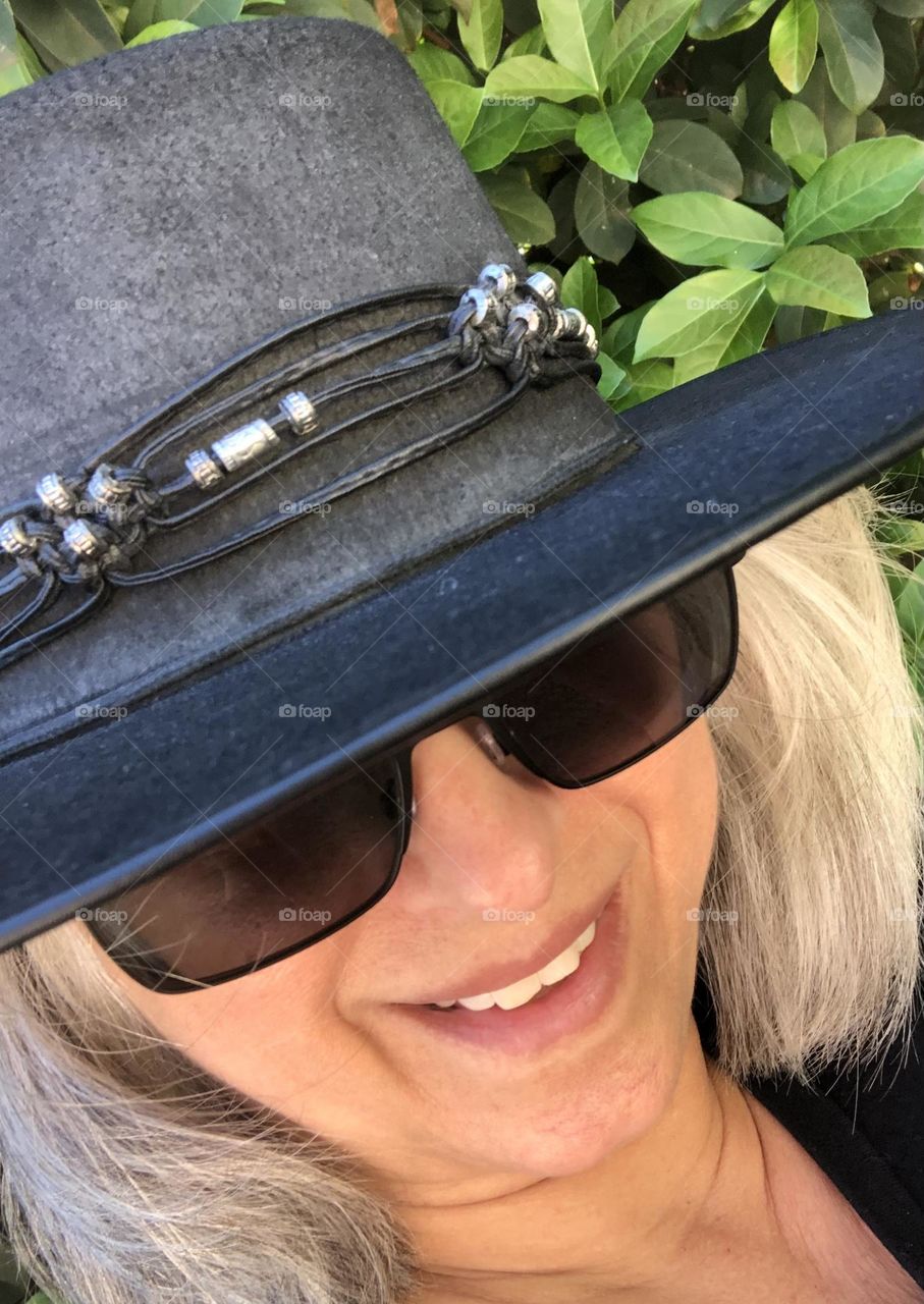 Closeup profile of smiling woman with gray hair wearing black hat and dark sunglasses 