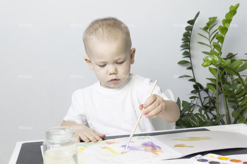 A small child draws with watercolors, multi-colored paints in an album at the table.