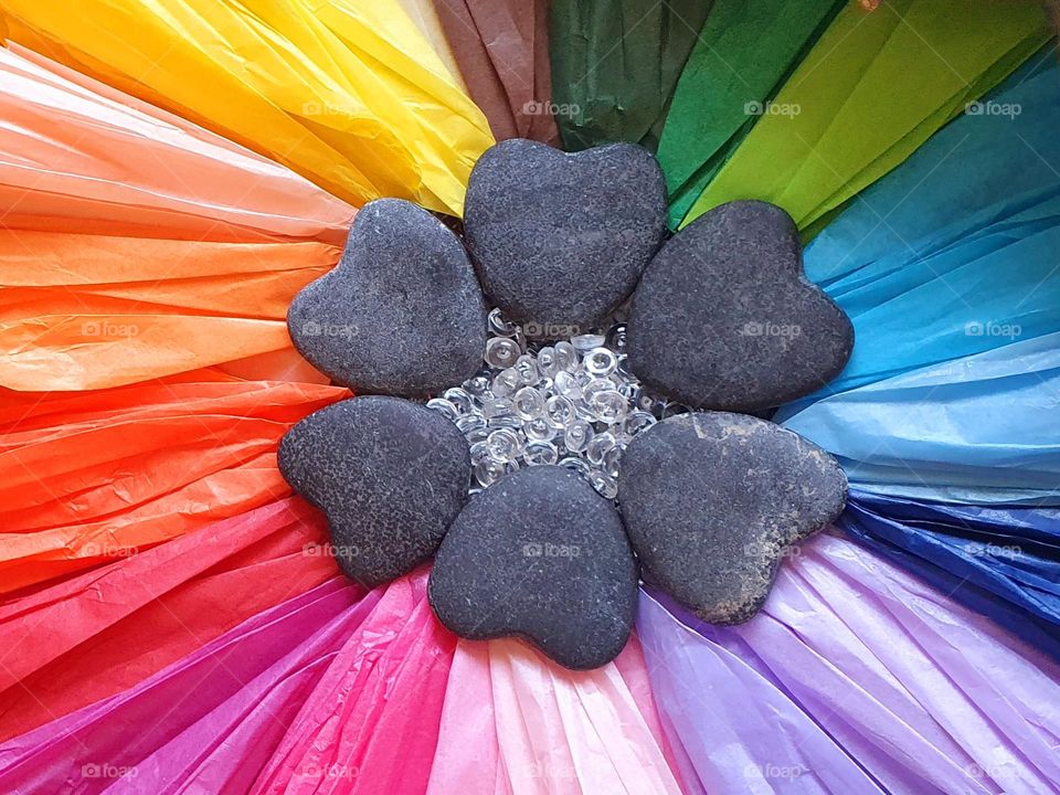 Flatlay composition of a flower's heart made of multi-coloured silk papers, heart-shaped pebbles and clear beads, creating a colour explosion