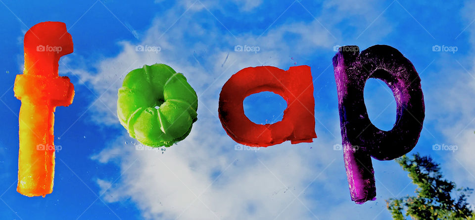 The foap name spelled out in jello letters and placed on a mirror outside on a sunny, partially cloudy, day.  There is a reflection of the clouds and a tip of a tree and the letters look like they’re floating in the sky!
