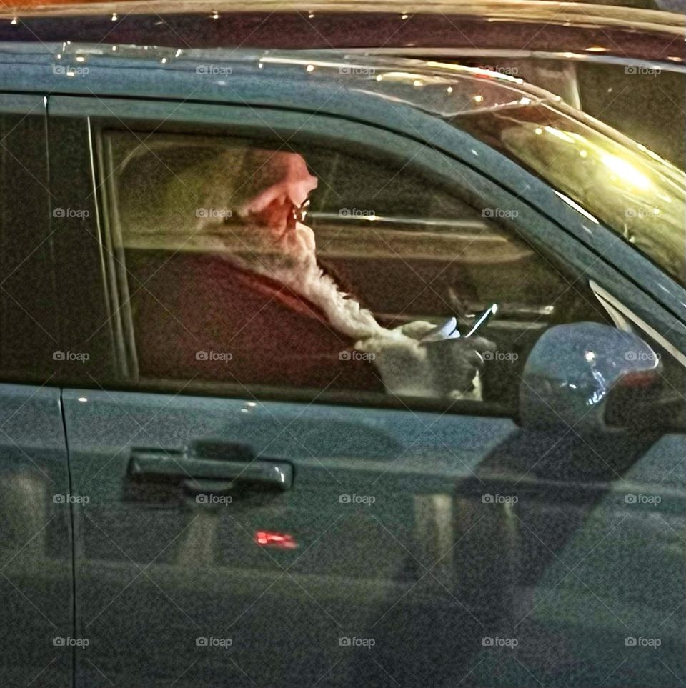 Santa sitting in a car in the driver’s seat checking his list on a cellphone while stopped at a traffic light