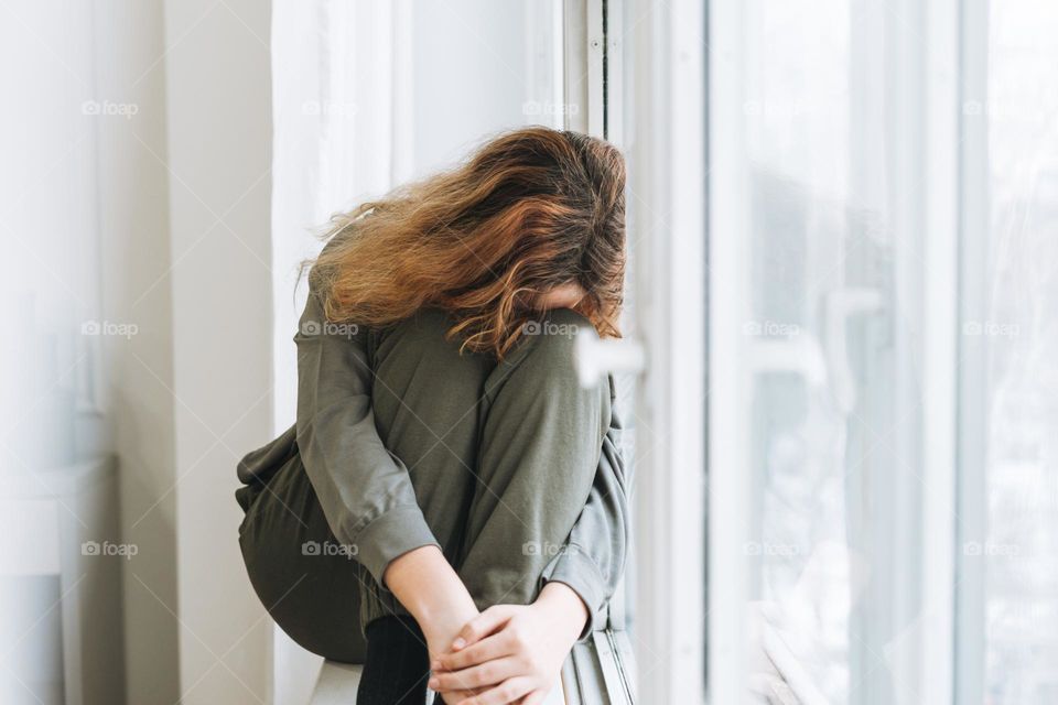 Beautiful sad unhappy teenager girl with curly hair in close pose sitting on window sill