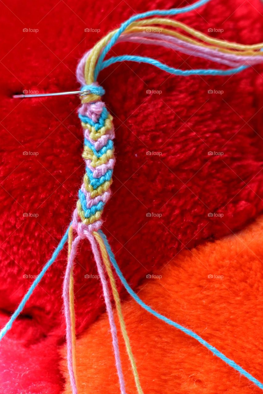 A homemade, colorful, knotted friendship bracelet hangs on a red pillow to be completed