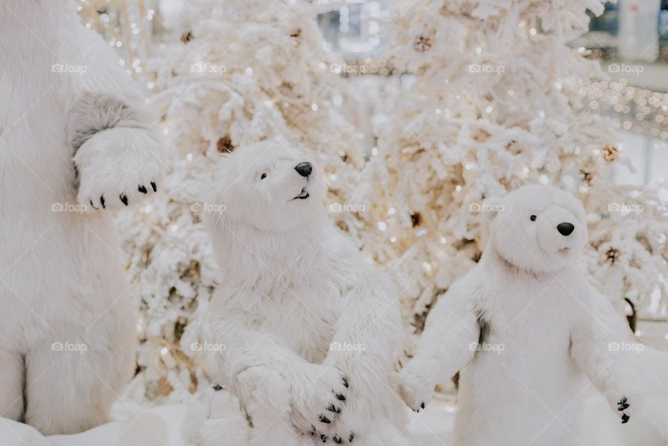 Large fluffy white toy bears on the background of white Christmas trees with burning garlands in the shopping center, close-up side view. Concept white on white, toy bears.