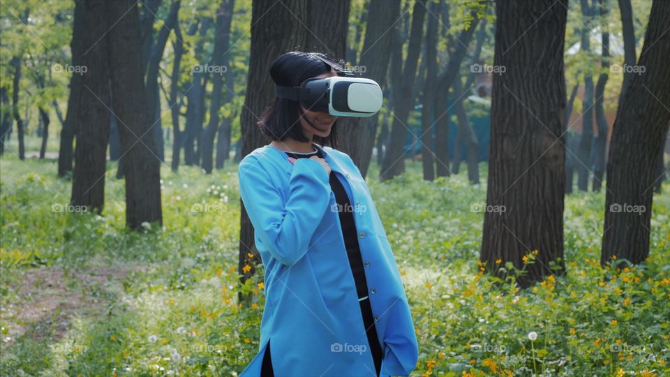 Portrait of trendy woman in blue coat using virtual reality glasses simulator in the park or forest at spring 