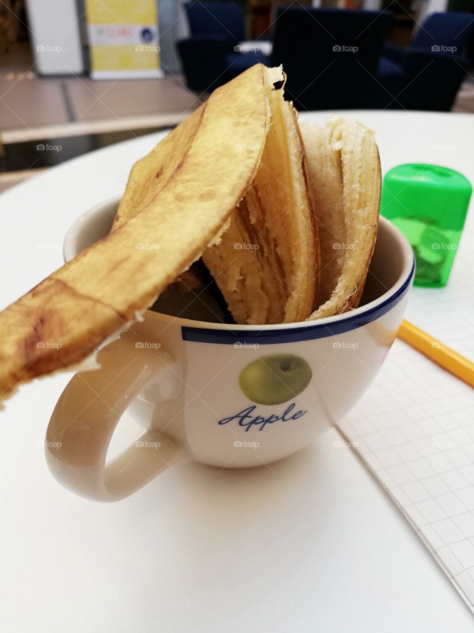 A banana skin in a coffee cup. A drawing of an apple on the surface. A pencil on the sheet of a paper and a green sharpener behind the mug on the table.