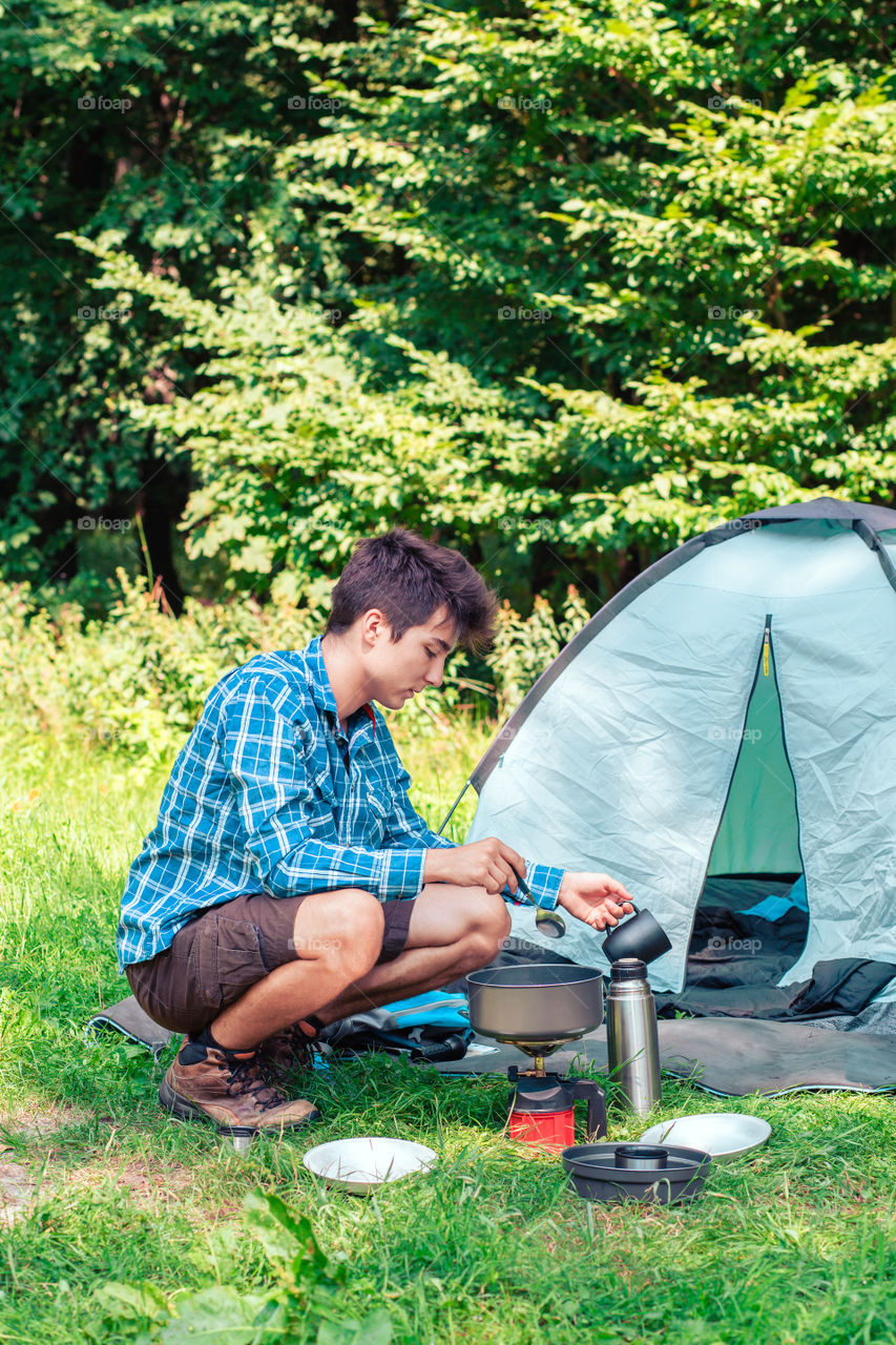 Spending a vacation on camping. Young man preparing a meal outdoor next to tent