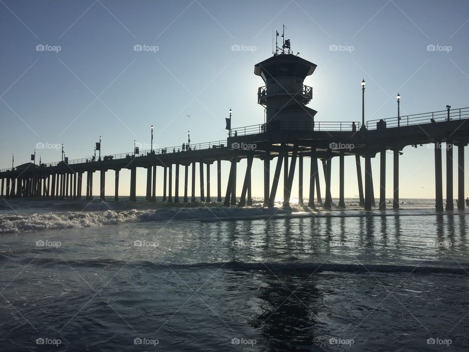On a glorious summer day, the pier at Huntington Beach, California glows from the light of the sun behind it, as the waves begin their retreat back to the ocean, readying themselves to crash onto the shore once again. 