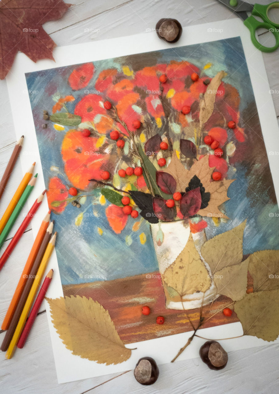 Children's creativity. Crafts, children's painting, applique with autumn leaves and mountain ash on a light wooden background with chestnuts, multi-colored dry leaves and pencils. Vase of flowers, dry autumn foxes, twigs and mountain ash