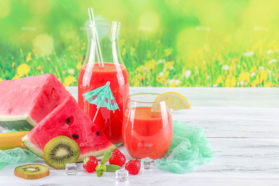 Multivitamin drink in a decanter and a glass with fresh fruits, berries: slices of kiwi, lemon and watermelon, mint leaves, rosemary and ice cubes on a light shabby table against a blurred window with a flowering field, close-up side view. Concept su