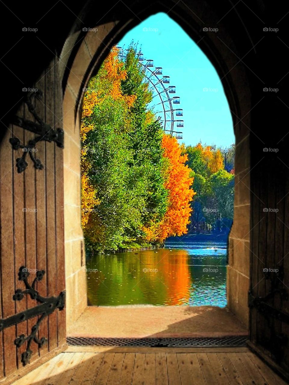 Autumn.  An open wooden door through which an autumn landscape opens.  The blue water reflects the colorful autumn trees.  Part of the ferris wheel is visible behind the trees