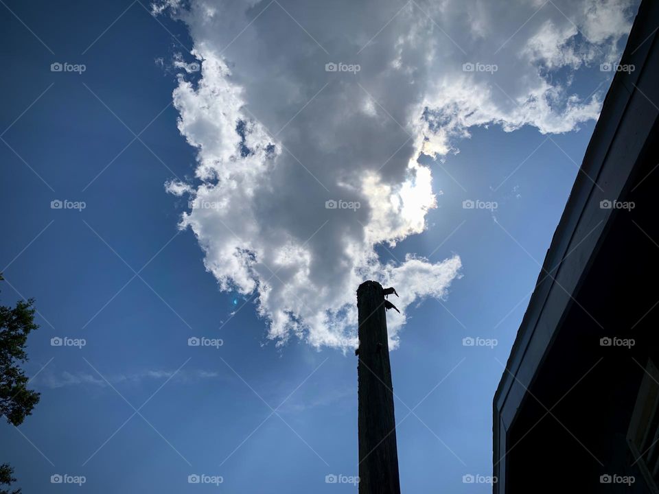 Cumulus Cloud In Blue Sunny Sky Looking Like Smoke Coming Out From The Pole By The House With Shadows And Rays In The Sky And The Sun Hiding Behind The Cloud.