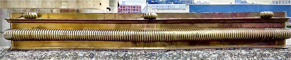 This rare found object of old fashioned interior design fit for a mansion or a palace, a fireplace fender made from bronze is photographed against the Greenpoint, Bklyn side of Newtown Creek in LIC, Queens, NY. 2022. Hypnotic Productions