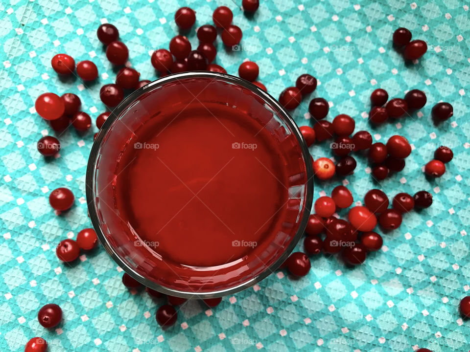 Many red ripe cranberries nearby a glass of fresh cranberry juice on blue textile background 