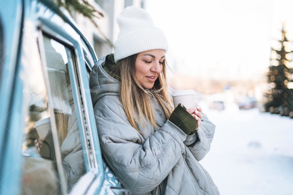 Young woman blonde girl in warm clothes with cup of coffee in hands near retro car decorated Christmas tree in winter street