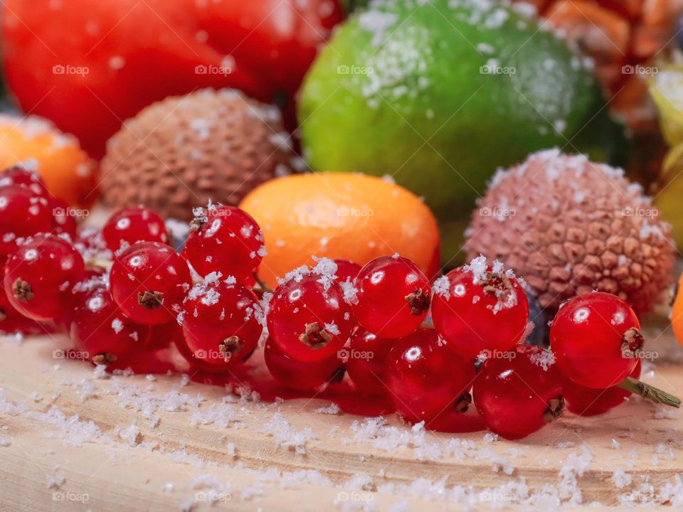 Branch of red currant with mini tangerine, lychee and persimmon with powdered snow on a wooden round dish, close-up side view. Holiday concept berries and fruits,healthy food