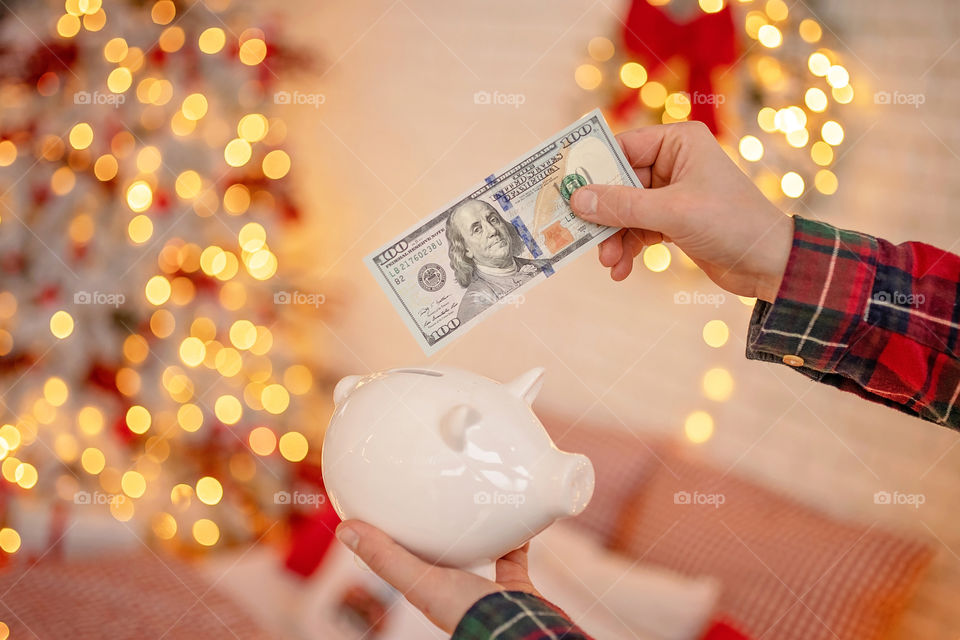 piggy bank with 100 dollars banknote in festive New Year atmosphere of scenery.  Magical bokeh with Christmas tree and bright lights.