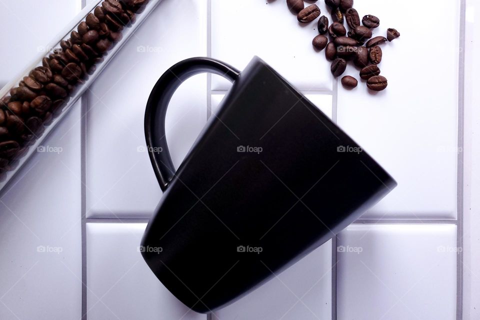 Coffee beans spilling out of slender glass container into a diagnolly tilted black coffee mug in front of a vertical white kitchen tile background.