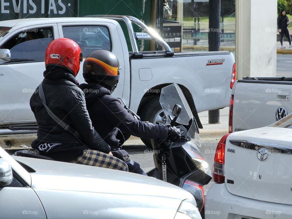 Two people on a motorcycle waiting at a light surrounded by cars on a busy street