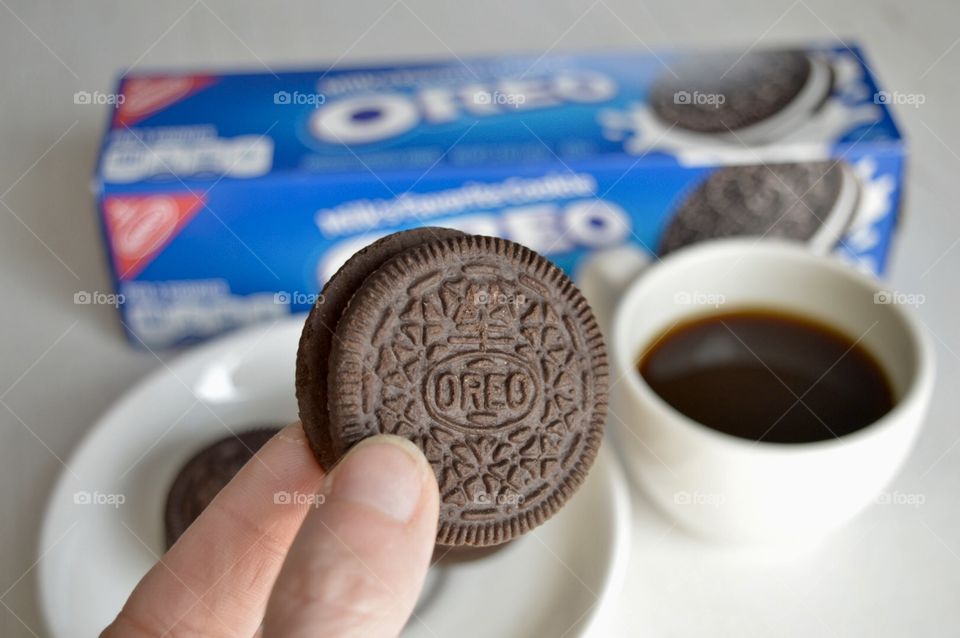 A persons fingers holding an Oreo cookie and a cup of coffee in the background