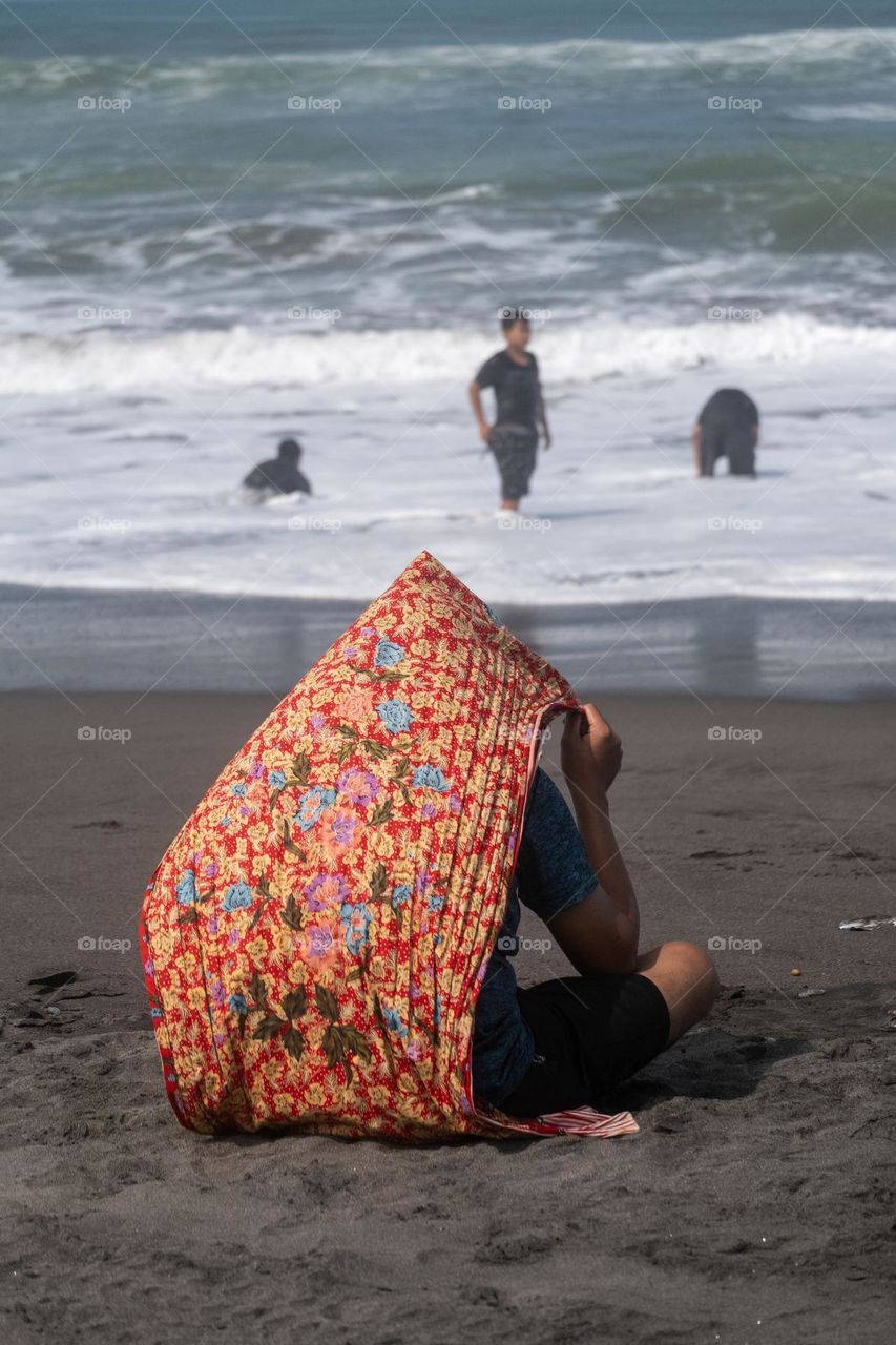 a man taking shelter from the hot sun by using a shawl on the beach