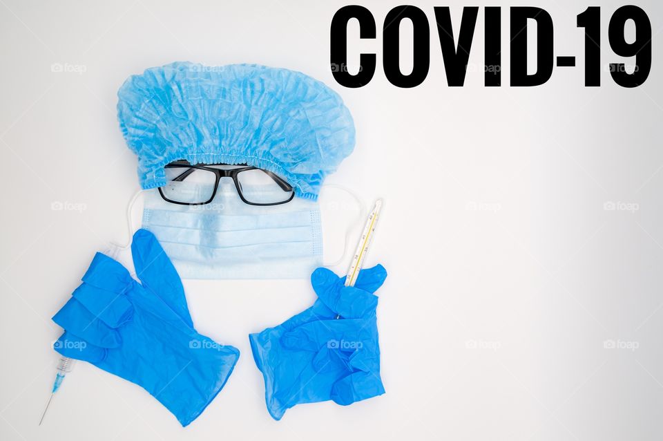 on a white background are medical objects laid out in the form of a face, a medical mask, rubber gloves, a thermometer, a cap, glasses and a syringe, all in blue. Top inscription "COVID-1