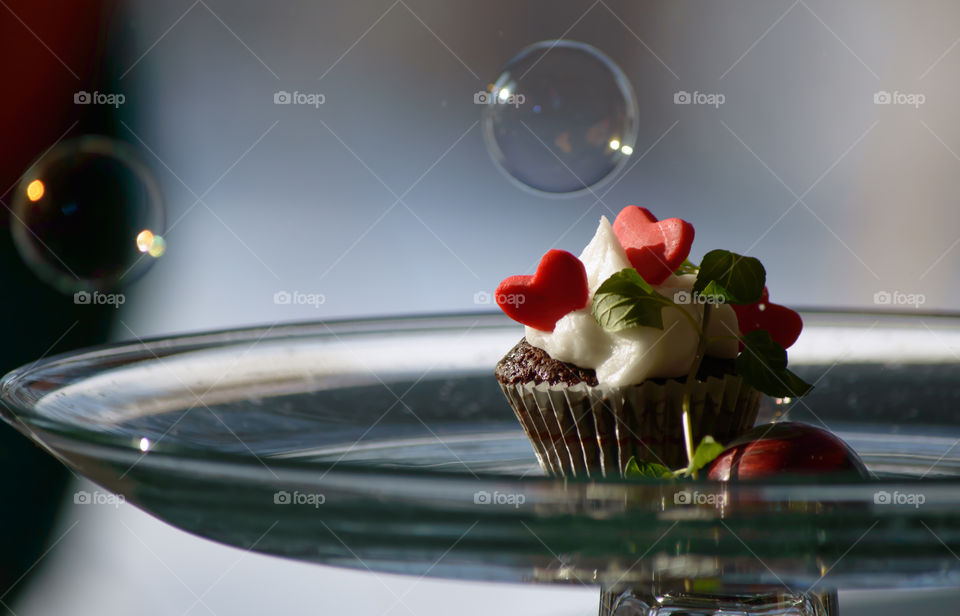 Chocolate cupcake with white buttercream frosting and candy hearts on glass pedestal plate with floating bubbles conceptual dreamlike love or party sweet food photography 