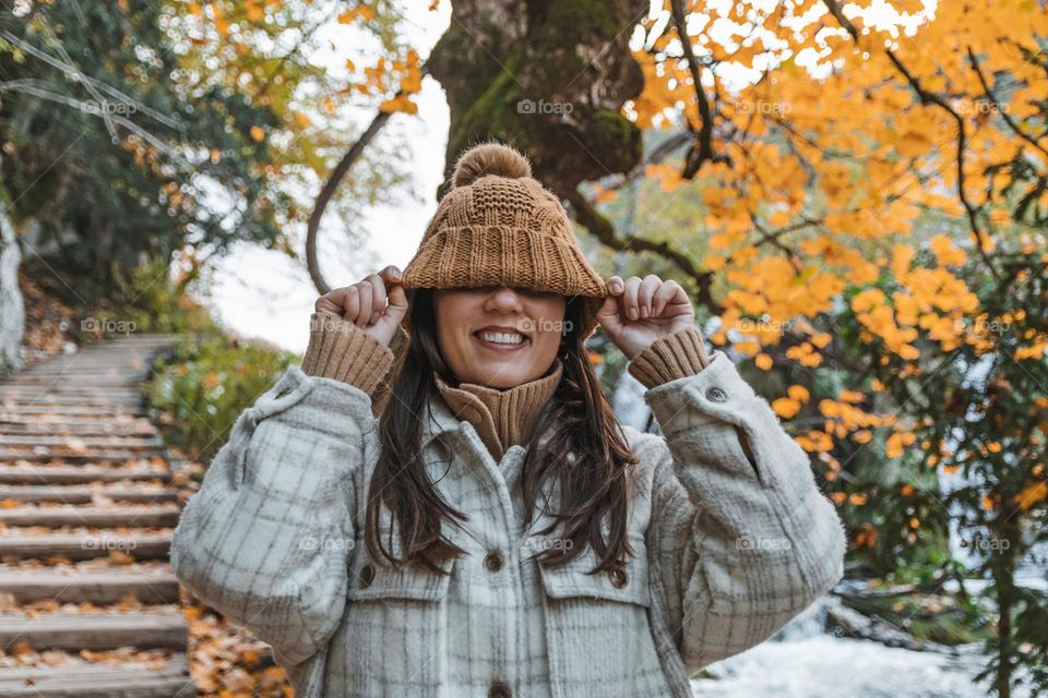 Portrait of happy girl standing on wooden footpath in forest in autumn. Young woman pulling knitted hat down over her eyes.