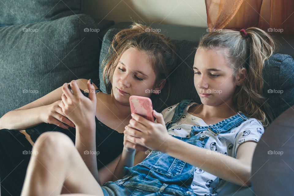 Young women using mobile phones watching music clip, texting, messaging. Teenagers using the smartphones, sitting on sofa at home. Using technology devices. Girls wearing summer clothing